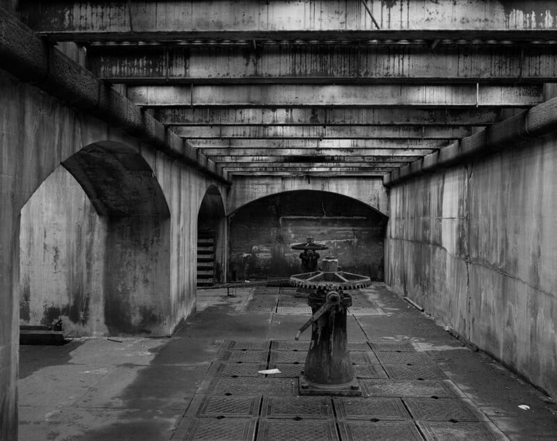 Black and white photo of an industrial, concrete interior with large pipes overhead, exposed beams, and two large metal valve mechanisms on the ground, creating a gritty, abandoned atmosphere.