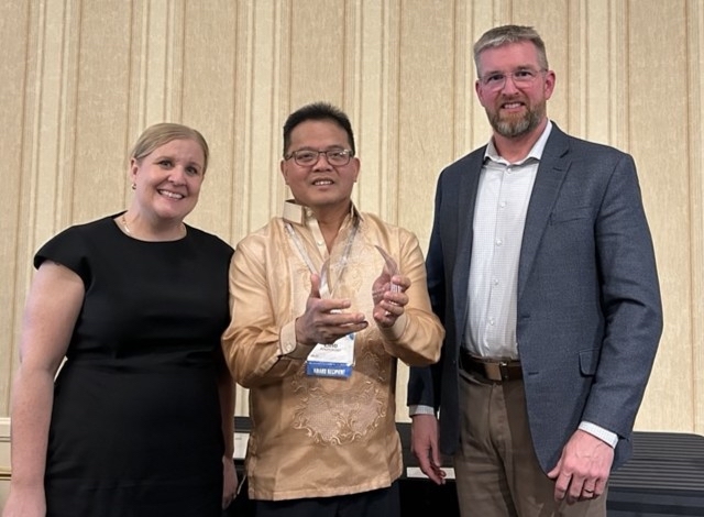 three people, with the man in the center holding up an award he just received