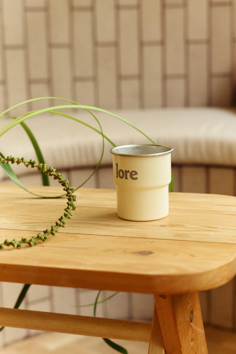 A cream-colored metal cup labeled "lore" sits on a wooden table, with a green plant partially visible in the foreground and a cushioned bench in the background.