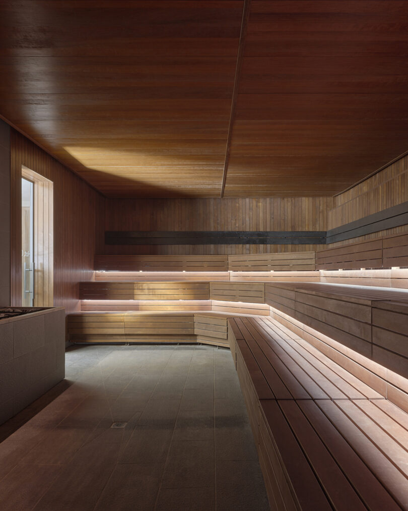 Modern wooden sauna room with tiered bench seating, natural light entering through a window, and stone flooring.