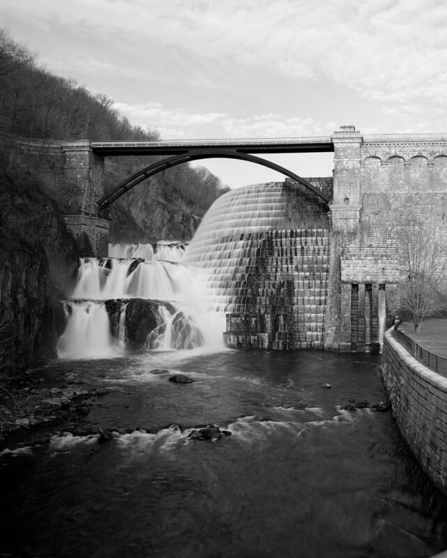 A stone dam with cascading waterfalls is spanned by two arched bridges, surrounded by trees and a cloudy sky, with water flowing into a river below.