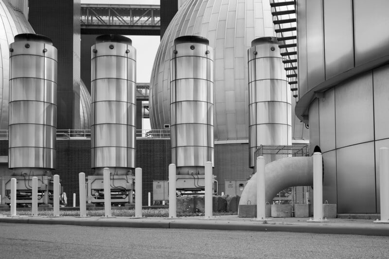 Four large cylindrical metal structures stand in front of round, dome-shaped industrial buildings, with pipes, fences, and vertical posts visible; the scene is in black and white.