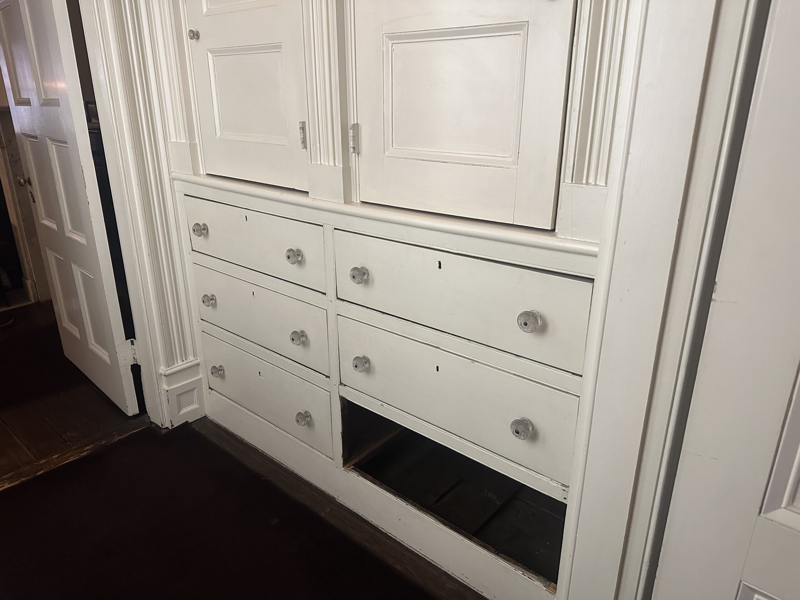 A series of white wooden drawers, some fitted with clear glass knobs, is shown on the wall. The top section includes cabinet doors, while the lower section has five drawers, with one drawer missing, revealing an open space underneath. A partially visible white door is seen on the left side, and dark carpet covers the floor. The overall setting suggests an older home with classic architectural details.