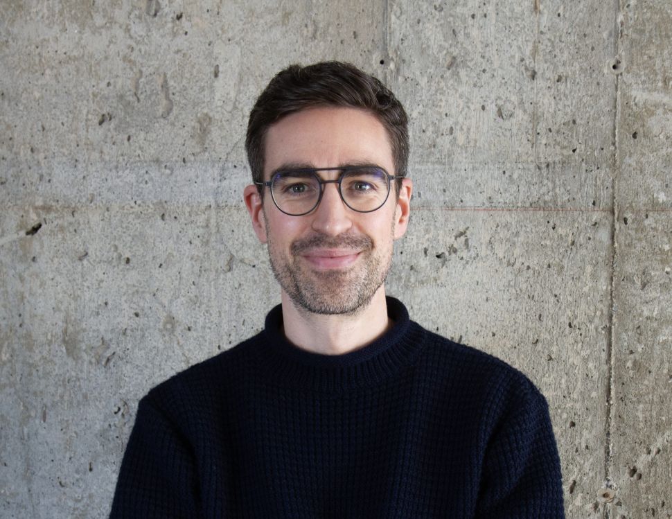 A man with short dark hair, wearing glasses and a navy sweater, stands with his arms crossed in front of a textured concrete wall, smiling at the camera.