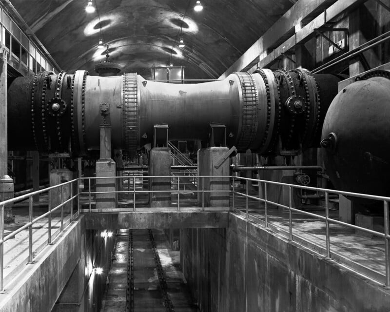 A large industrial tunnel or duct with gears and pipes inside a spacious, arched facility, viewed from a central walkway with railings. Overhead lights illuminate the concrete walls and metal machinery.