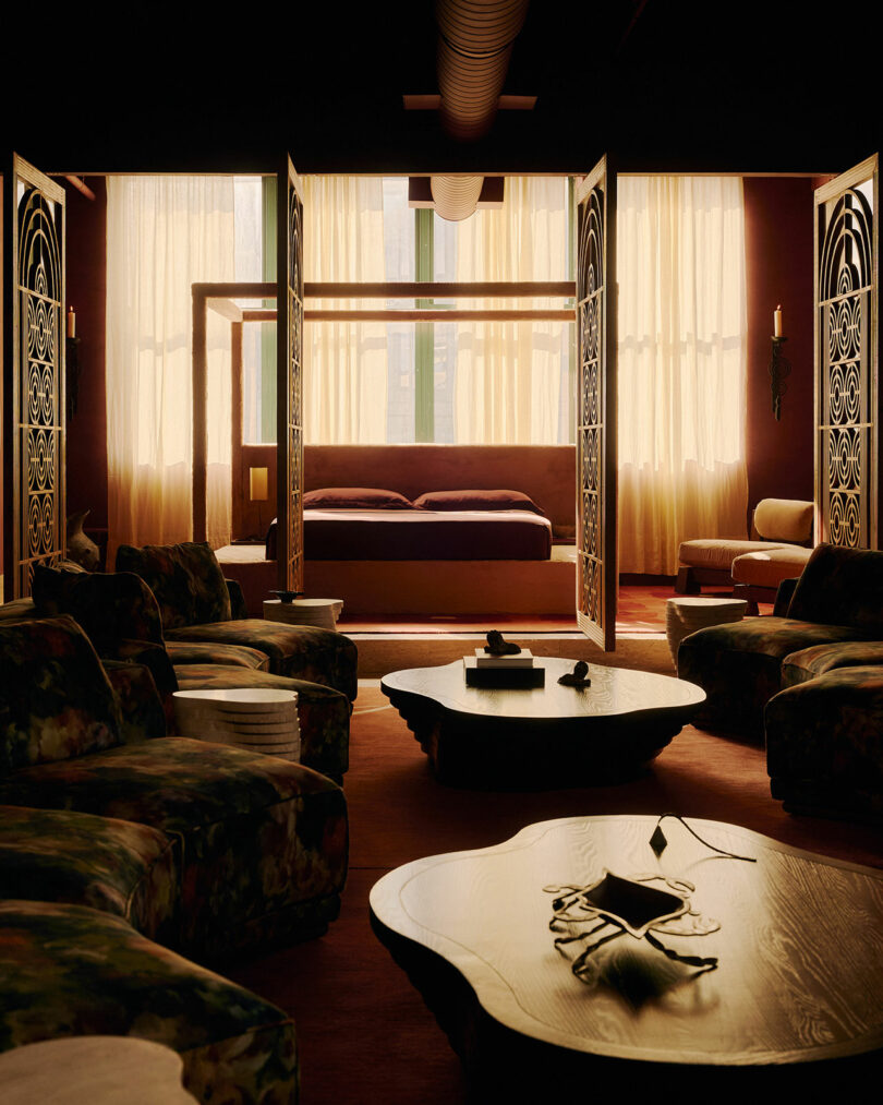 A dimly lit room with ornate folding screens, a canopy bed in the background, sculptural coffee tables, and plush patterned seating arranged in a circle. Soft light filters through sheer curtains.