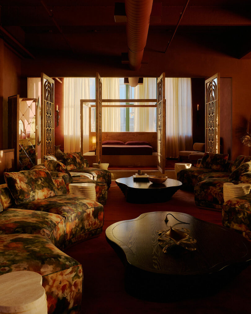 Dimly lit lounge with floral-patterned chairs, dark wooden tables, and a canopy bed in the background, separated by decorative screens and sheer curtains.