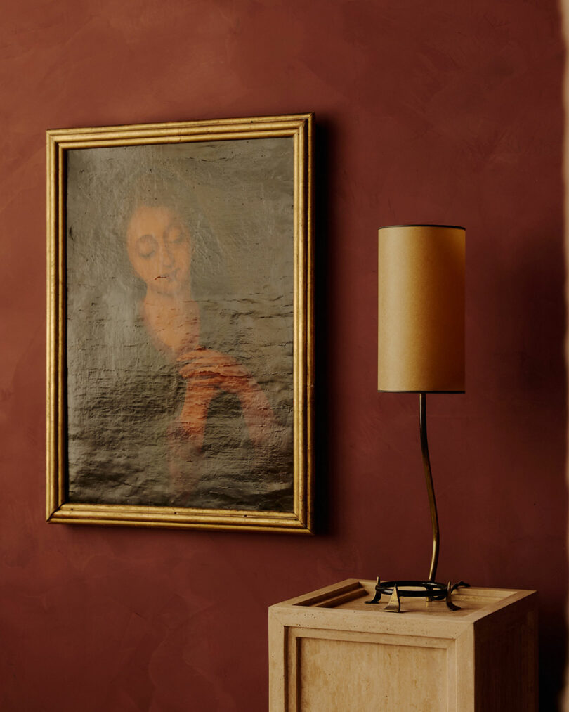 A framed, faded portrait painting hangs on a red wall above a beige pedestal with a modern, cylindrical beige lamp.