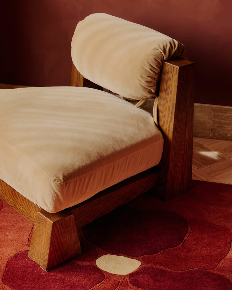 A wooden lounge chair with beige upholstered seat and backrest sits on a red floral-patterned rug against a reddish-brown wall.