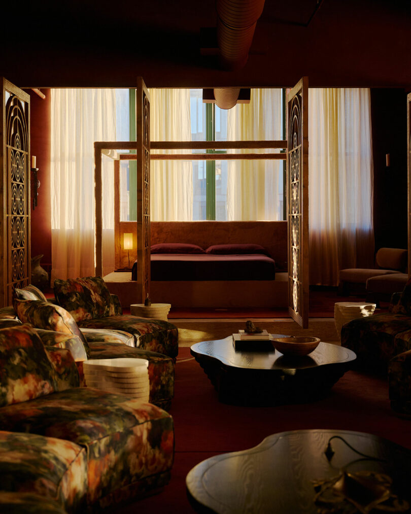 A dimly lit room features a canopy bed with decorative panels, floral armchairs, a sculptural coffee table, and sheer curtains covering tall windows.