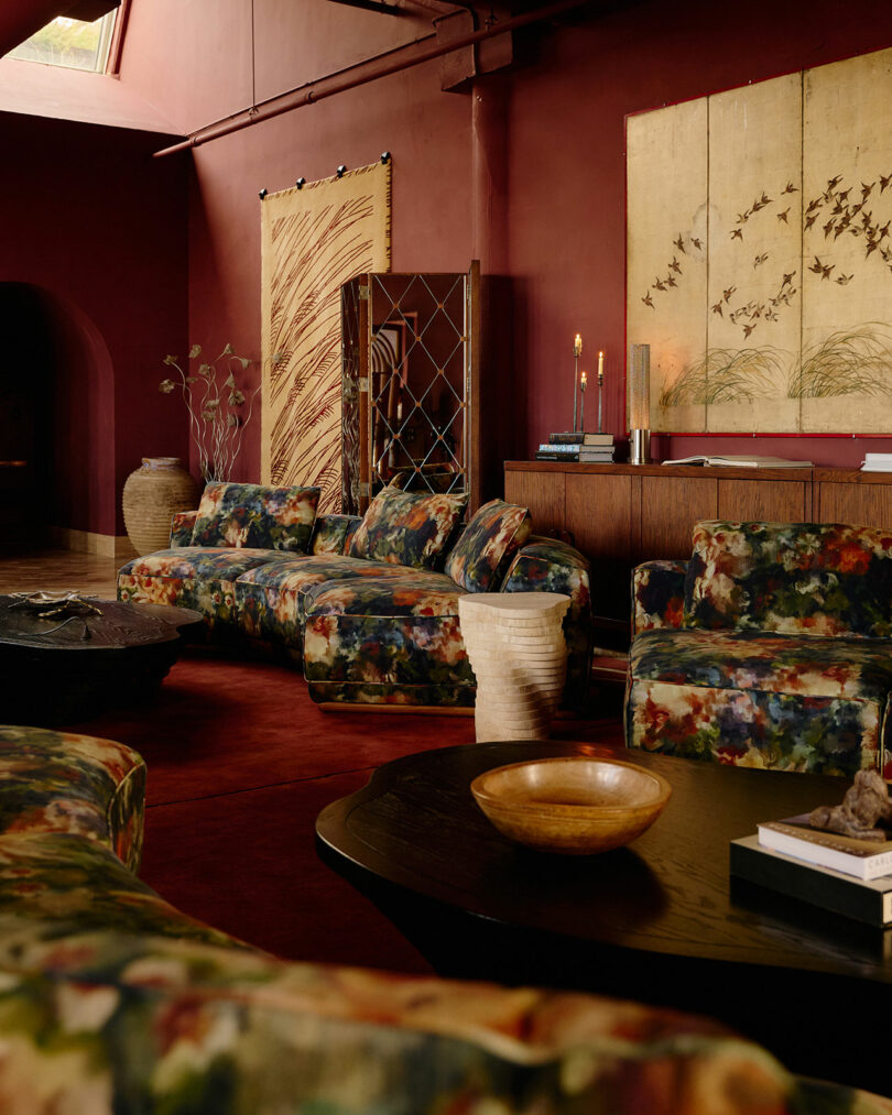 A living room with deep red walls, patterned floral sofas, a wooden coffee table, decorative screens, candles, and large wall art featuring birds and grasses.
