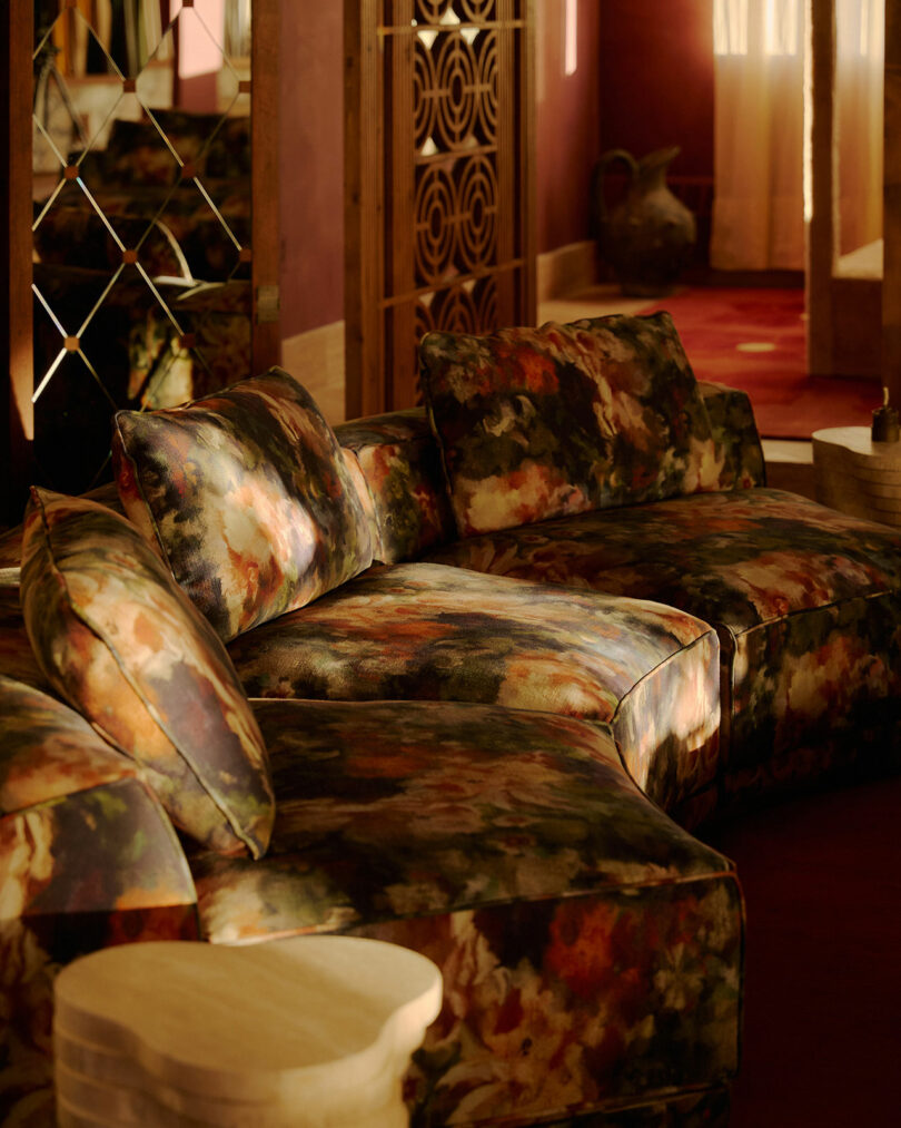A floral-patterned sectional sofa sits in a sunlit room with red walls, mirrored panels, and wooden accents.