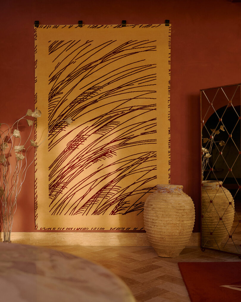 A large abstract-patterned rug hangs on a red wall above a parquet floor, with two textured vases and a mirrored cabinet nearby.