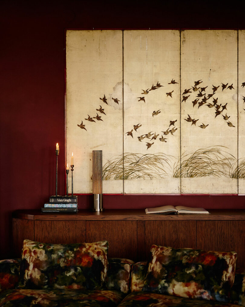 A wooden sideboard against a dark red wall with a large artwork of flying birds above, two candles, stacked books, a lamp, and an open book; a floral-patterned sofa is in front.