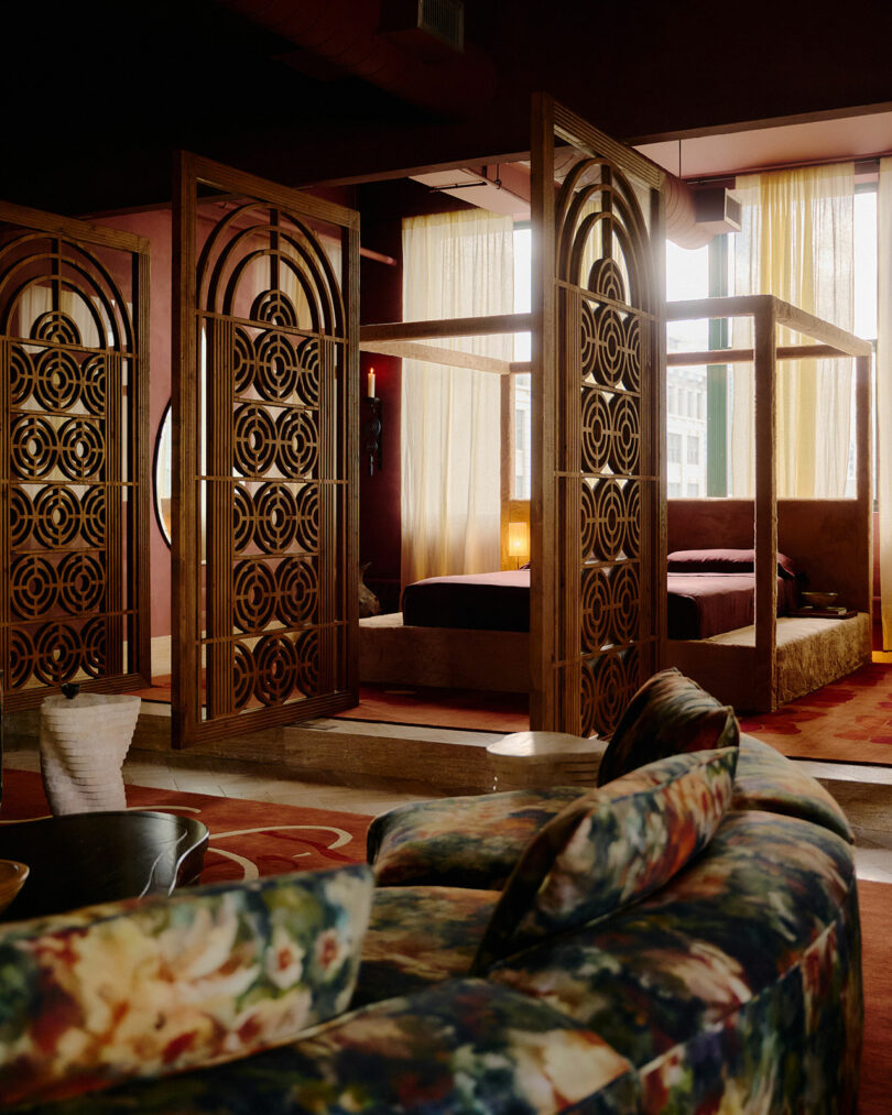 A stylish room features a floral-patterned sofa, a bed with a canopy frame, wooden decorative screens, and large windows with sheer curtains letting in natural light.