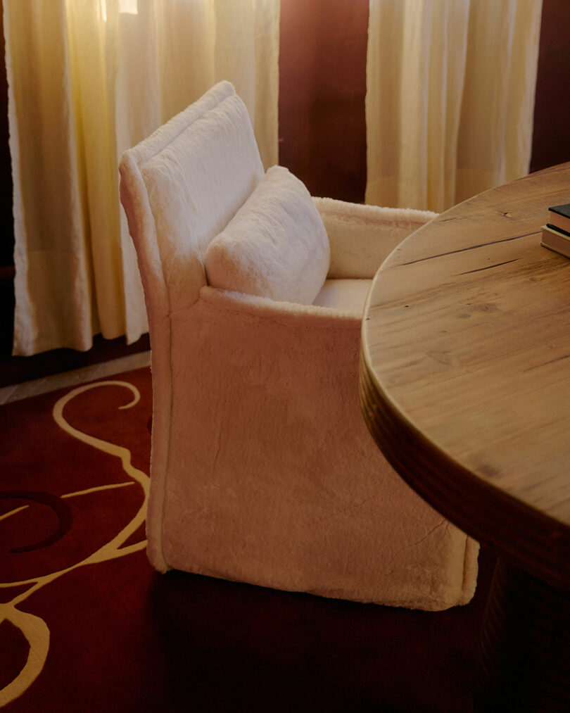 A white upholstered chair with a matching cushion is placed next to a wooden table, with yellow curtains and a red carpet in the background.