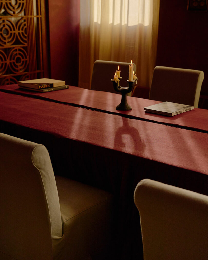 A dimly lit room with a maroon tablecloth, four white chairs, a candelabra with melting candles, and two books on the table near a window with sheer curtains.