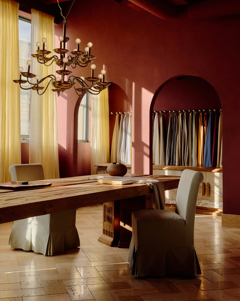 A sunlit room with red walls, a long wooden table, upholstered chairs, chandeliers, and fabric samples hanging on racks by arched windows with sheer curtains.