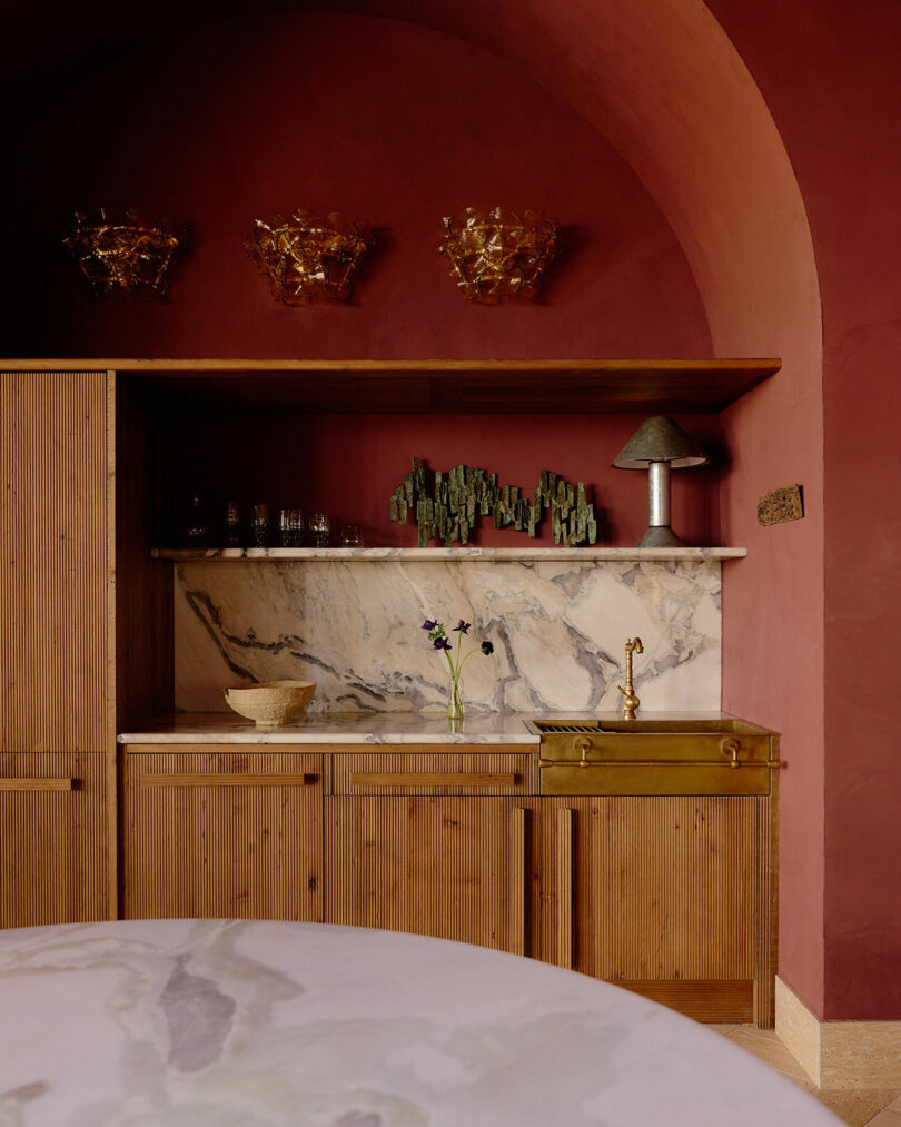 A kitchen with burgundy walls, wood cabinets, marble countertops and backsplash, a brass sink, and decorative glass, metal, and ceramic objects on the shelves.
