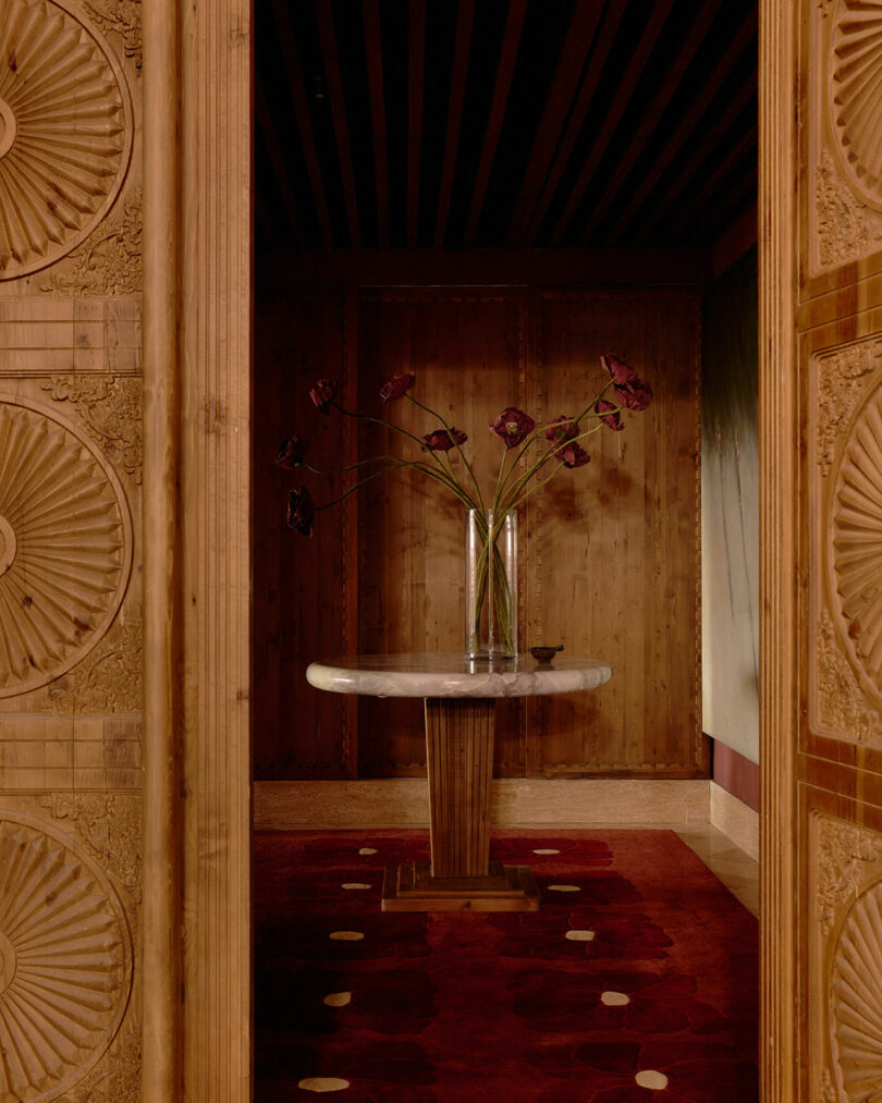 A small marble-topped table with a tall glass vase of dark red flowers stands on a red patterned rug in a wood-paneled room, viewed through ornately carved wooden double doors.