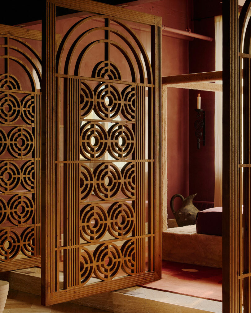 Wooden decorative room divider with geometric circular patterns stands in a warmly lit interior with earthy walls and a clay jug in the background.