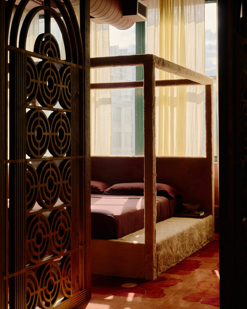 A sunlit bedroom with a four-poster bed, sheer curtains, geometric patterned screen, and red patterned carpet.