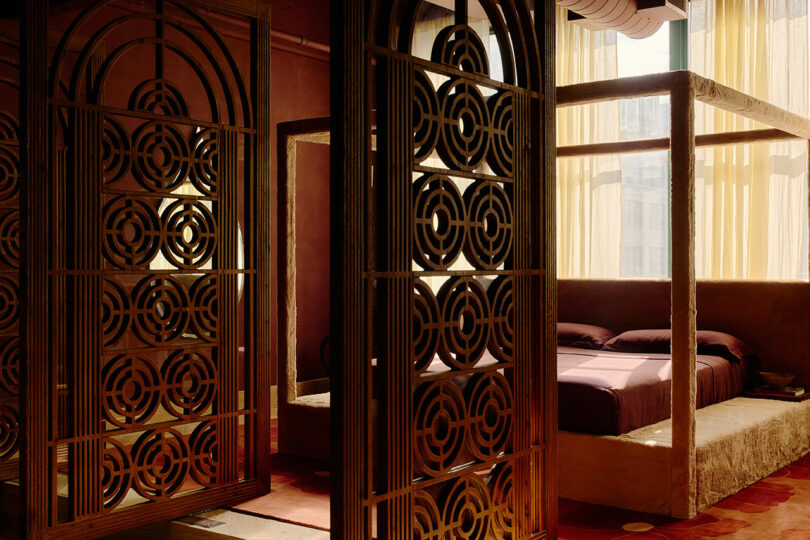 A room with decorative metal screens, a canopy bed with purple bedding, and yellow curtains filtering sunlight through a window.