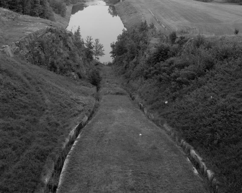 A grassy, sloped canal lined with vegetation leads to a calm body of water, surrounded by grassy fields and trees; the image is in black and white.