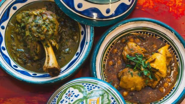 Overhead view of colorful Moroccan tagine dishes with lids. Lamb shank and chicken are served on a vibrant red table, evoking warmth and richness.