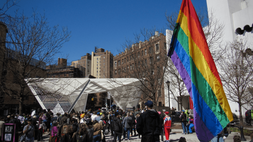 A person holds a large Pride flag outside of a cluster of people holding picket signs in front of the white New York City AIDS Memorial.