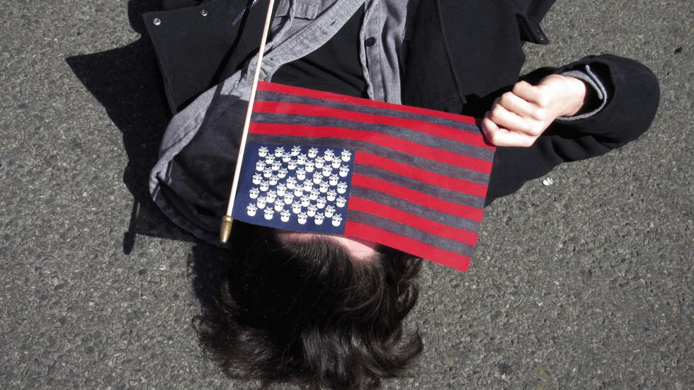 A person lies in the street and holds a black and red American flag over their face.