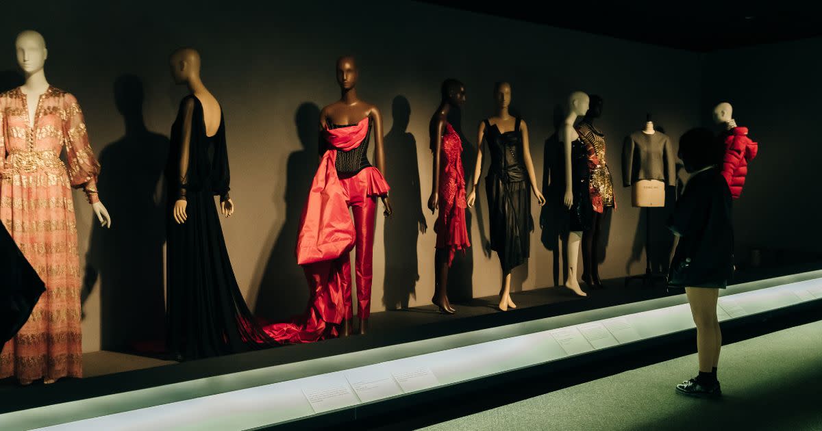 A fashionable exhibit at New York City's Fashion Institute of Technology (FIT). Stylish wardrobe and mannequins line a wall at a show exhibit, as an onlooker observes.