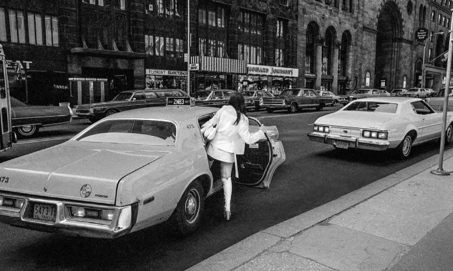 A woman in a white coat and boots steps into the back seat of a classic taxi on a busy city street, lined with parked cars and historic buildings in the background. The photo is in black and white.