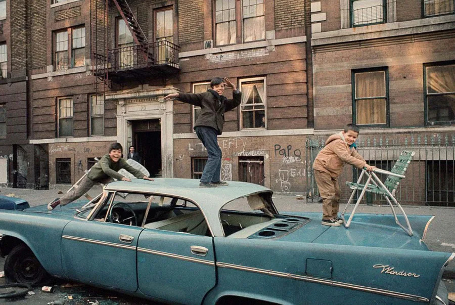 Three children play on top of a damaged blue car parked on a city street, with an old brick apartment building and graffiti in the background. One boy holds a chair, while others pose energetically on the car roof.