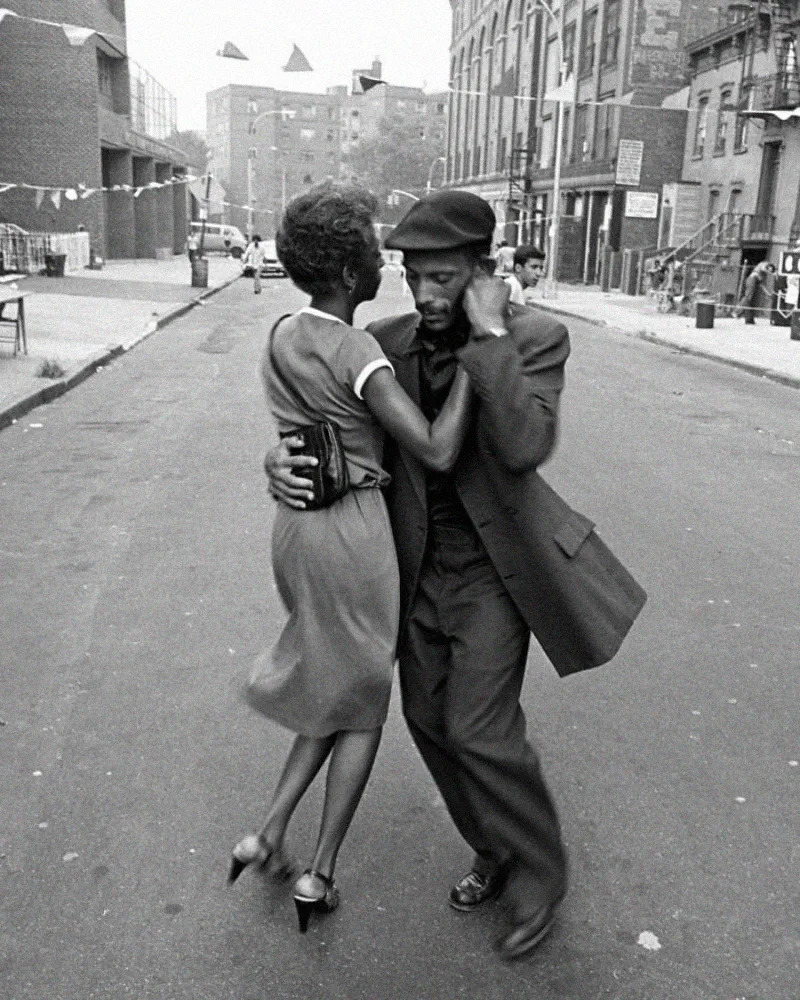 A man in a suit and hat and a woman in a dress dance closely together in the middle of an empty city street, with buildings and bunting visible in the background.