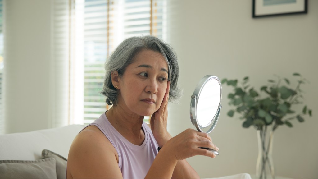 A middle-aged woman with gray hair looks into a mirror, touching her face and neck, with a stressed expression.