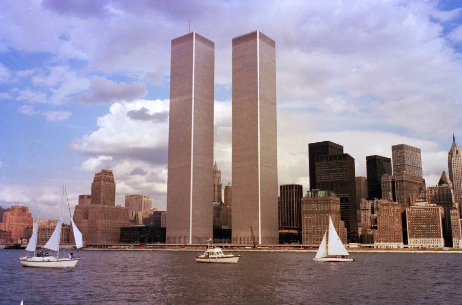 The World Trade Center’s twin towers rise above the Manhattan skyline in New York City, with several sailboats and a yacht on the water in the foreground under a partly cloudy sky.