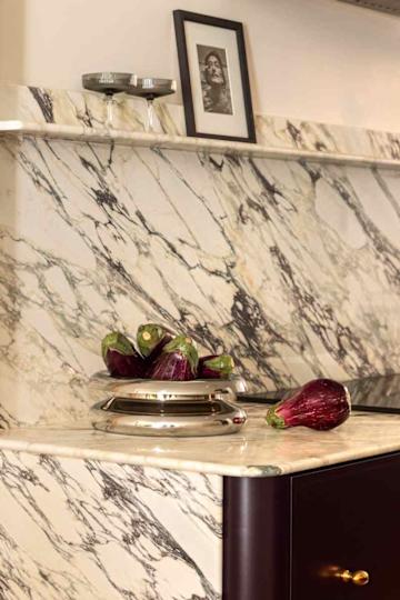 A polished marble countertop with a silver bowl holding several eggplants, framed photo, and two glass bowls on a shelf.