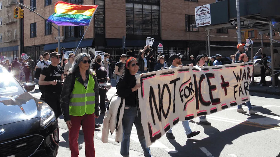 People holding a banner that reads "Not for ICE and War Fare" walk through a city street.
