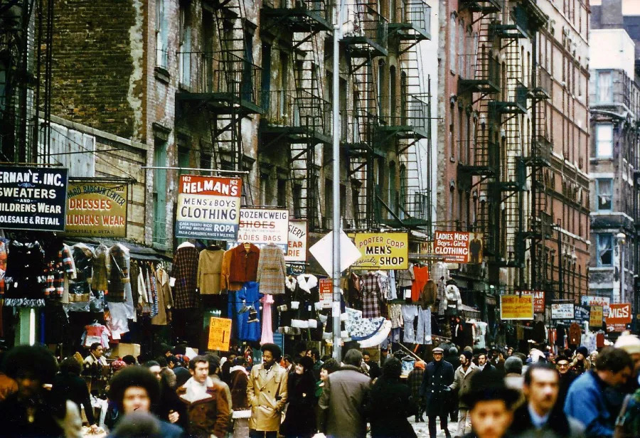 A busy city street lined with old brick buildings, fire escapes, and many colorful signs advertising clothing shops. Crowds of people walk past outdoor displays of hanging clothes and jackets on racks.