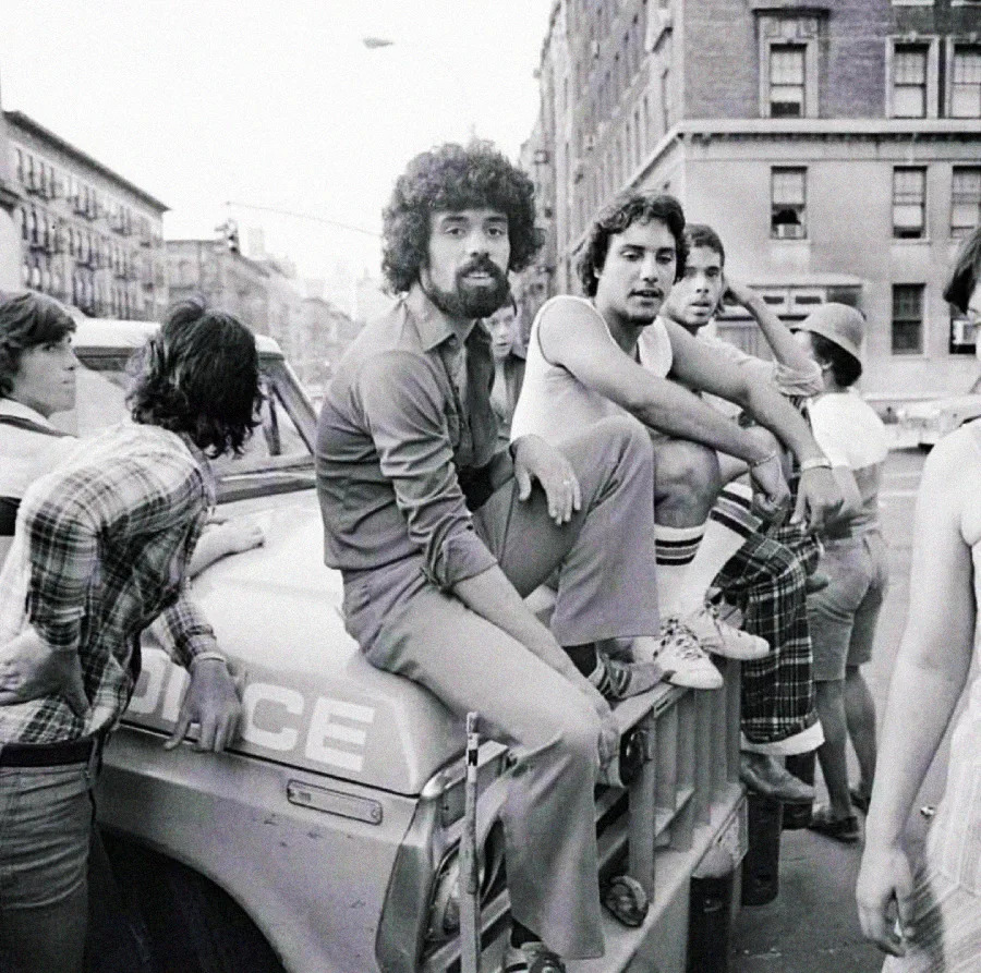 A group of young adults sit and lean on the hood of a police car parked on a city street, surrounded by tall buildings, in a black-and-white photo.