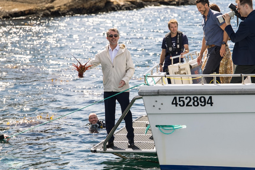 Danish King Frederik holds a lobster and smiles while he stands on the back of a boat.