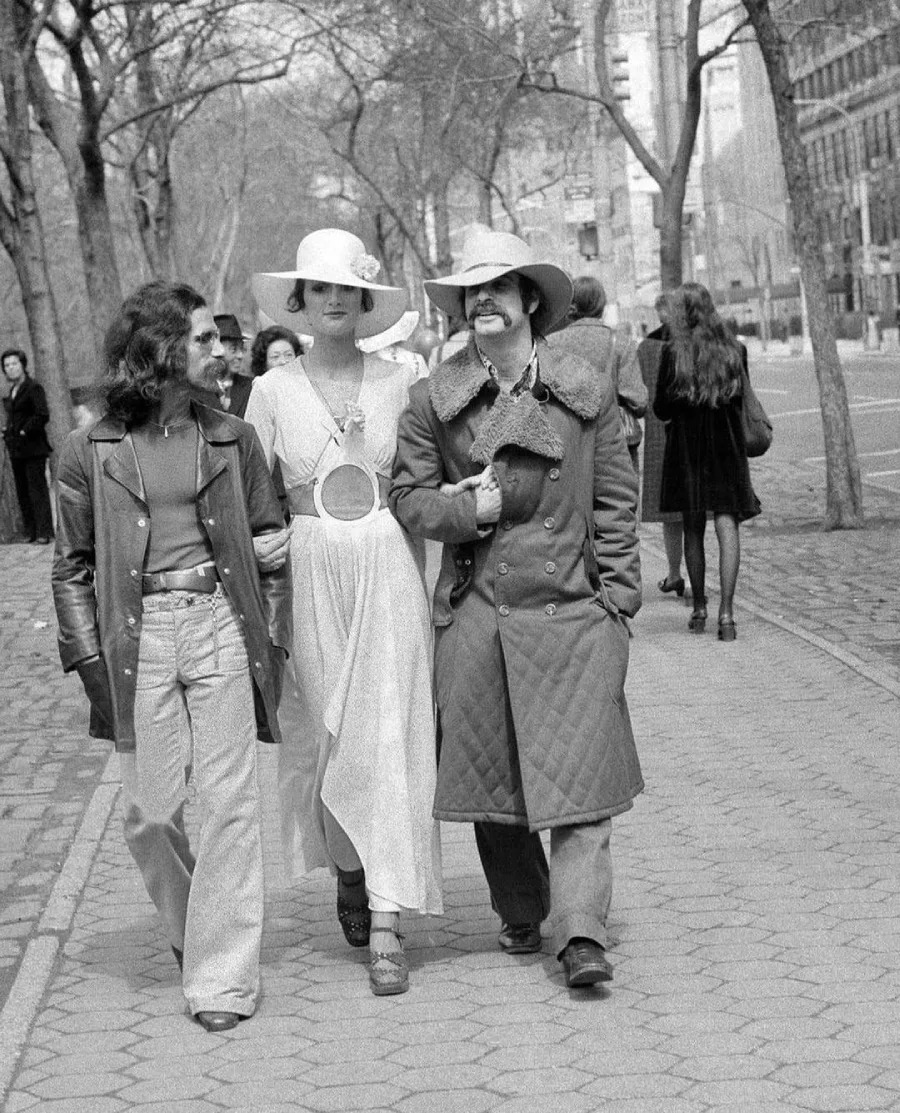Three people in fashionable 1970s outfits walk together down a city sidewalk, with trees and buildings in the background. The two men wear coats and hats, and the woman wears a white dress and wide-brimmed hat.