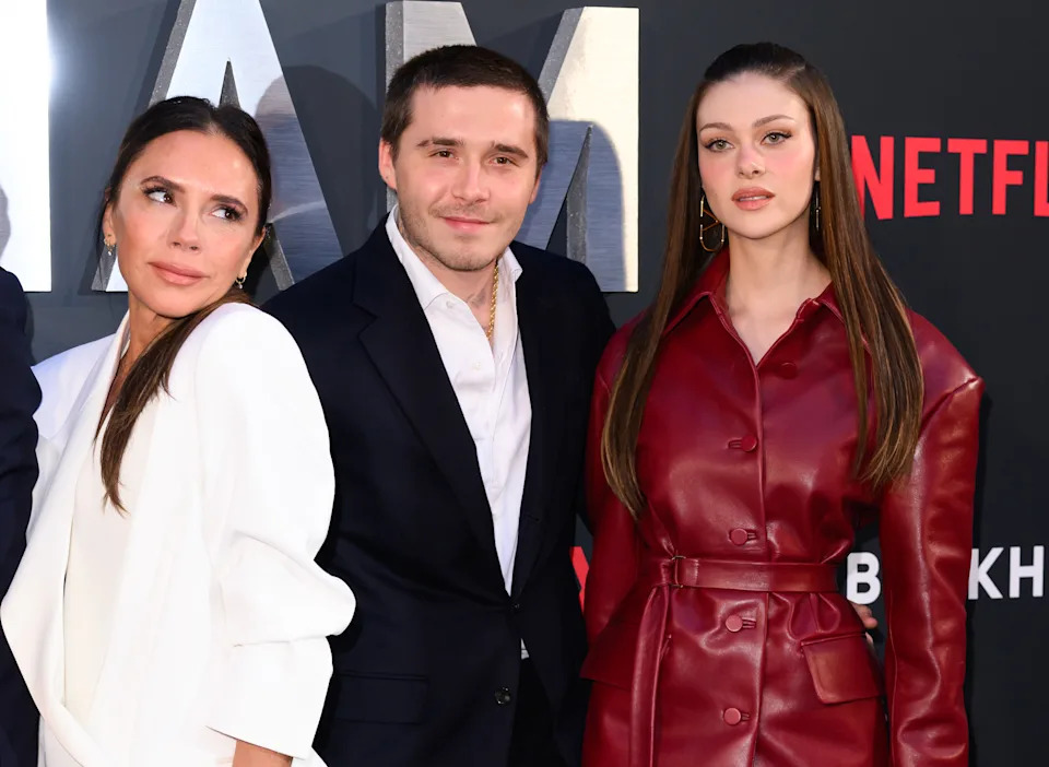Victoria Beckham in an elegant blazer and pants stands next to Brooklyn Beckham in a suit and Nicola Peltz Beckham in a long leather coat on a Netflix red carpet