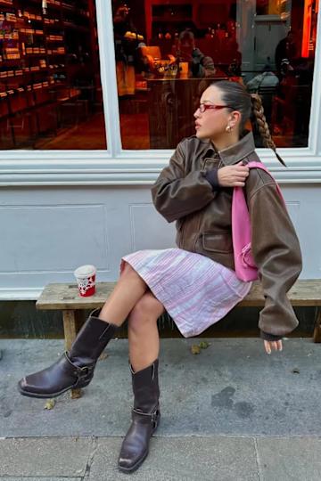 a woman wearing a brown leather jacket, pink shoulder bag, pink striped skirt, and brown boots