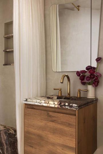 Modern bathroom with a wooden vanity, marble countertop, gold faucets, a mirror, and a vase of purple flowers.