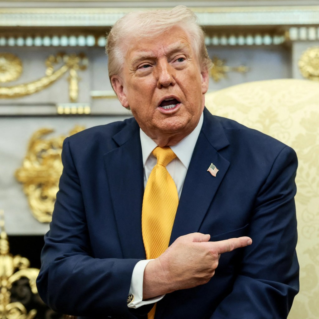 President Trump, wearing a dark blue suit and yellow tie, speaks and points in the Oval Office.