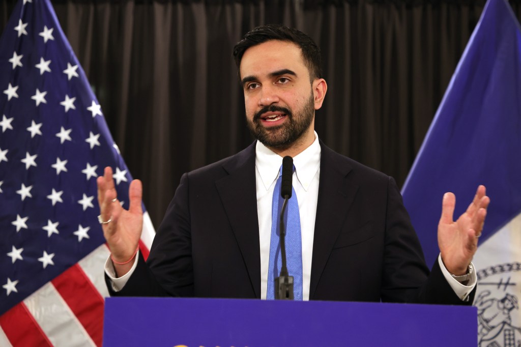 Mayor Zohran Mamdani speaking at a press conference with American and New York flags in the background.