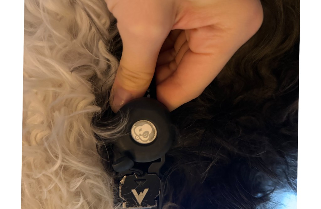 A hand holding a black tracking device with a cartoon face logo, positioned on the fur of a dog.