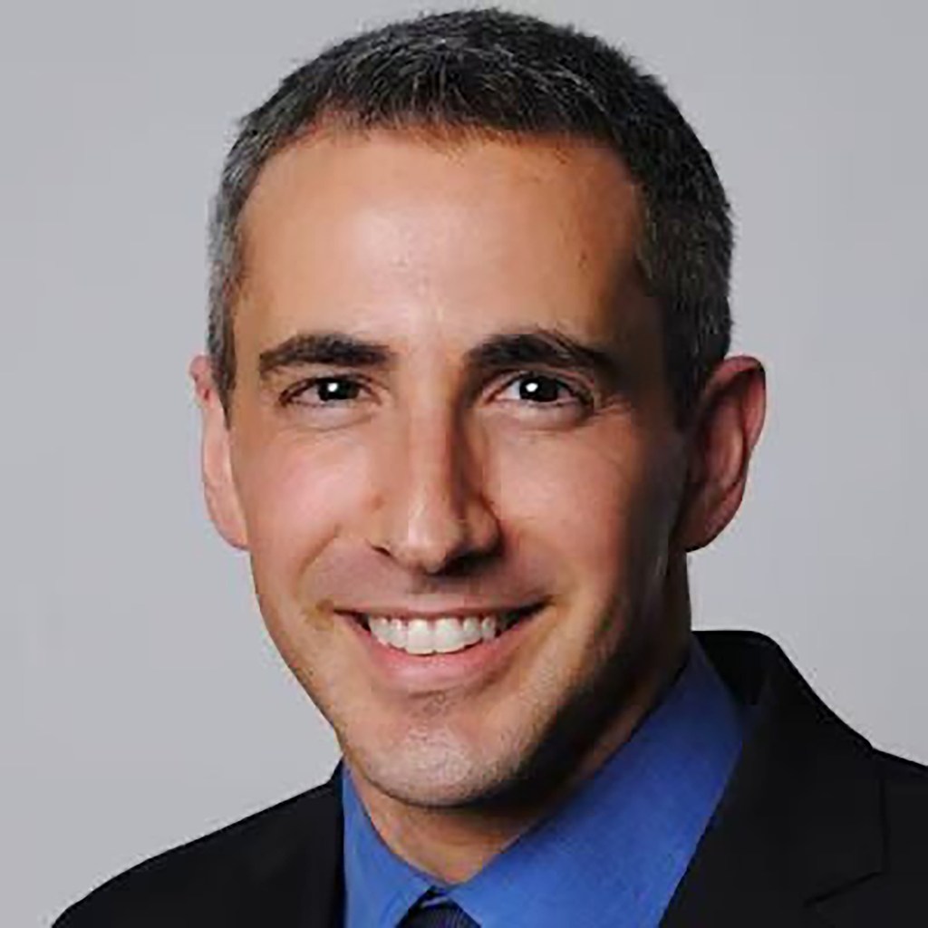 Dr. Tal Ben-Zvi smiling in a headshot against a gray background.